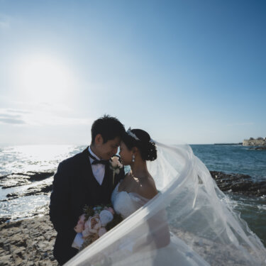 Bride and groom touching foreheads on the rocky shore as her veil flows in the sea breeze, beach wedding photography in Aichi, Japan