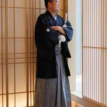 Groom in montsuki hakama leans against a lattice wall with his arms crossed, a relaxed solo shot from a Japanese wedding photoshoot
