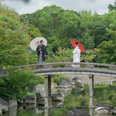 Wide shot of the groom with a white wagasa and the bride with a red wagasa standing apart on an arched bridge over the pond at Daisen Park Japanese Garden, a picturesque pre-wedding photoshoot in Osaka
