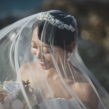 Smiling bride beneath a sheer veil with a soft pink bouquet, a dreamy bridal portrait from a seaside wedding photoshoot