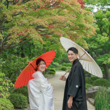 Bride in shiromuku with a red wagasa umbrella and groom in montsuki with a white one smiling back at the camera under maple leaves turning red, kimono wedding photos in a Japanese garden