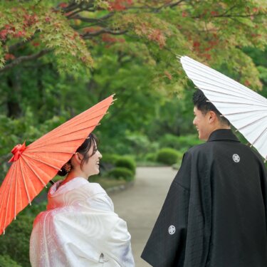 Bride with a red umbrella and groom with a white umbrella facing each other and laughing on a garden path under early autumn maples, a colorful pose idea for wedding photography in Japan