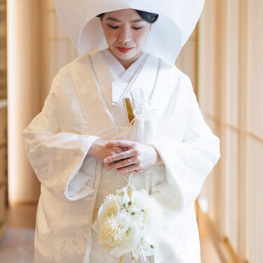 Bride in a round wataboshi hood and shiromuku looks down at her white flower bouquet, a serene kimono wedding photography portrait