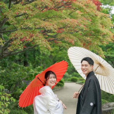 Couple in shiromuku and montsuki holding red and white wagasa umbrellas and glancing back with smiles beneath red-tinged maple trees in Daisen Park, a pre-wedding photo in Osaka