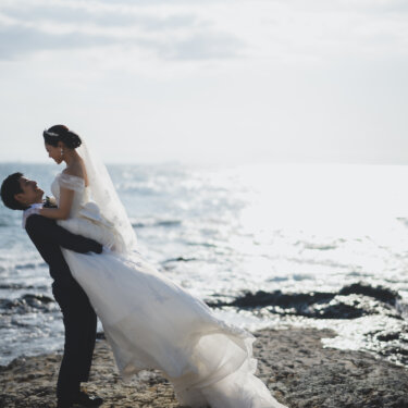 Groom lifting the bride high on the rocky shoreline with the sparkling sea behind them, a dynamic pose for Japan wedding photography