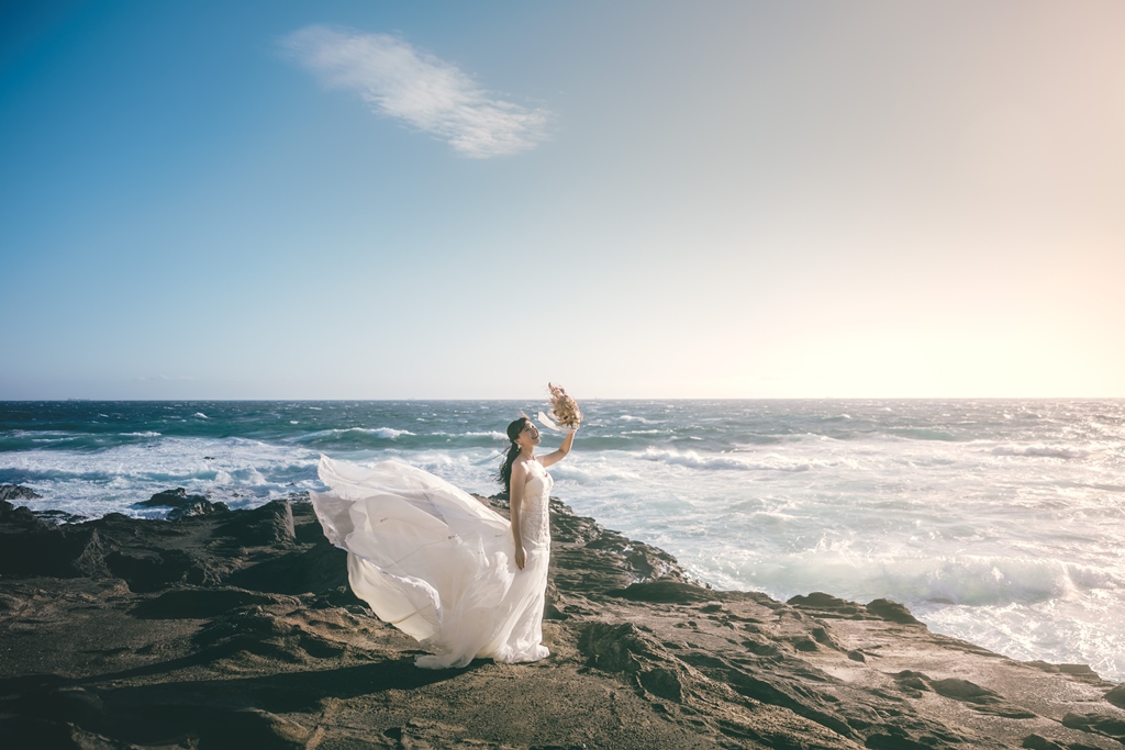 Bride's wedding dress and veil flowing in the sea breeze as the couple stands on the rocks of Jogashima