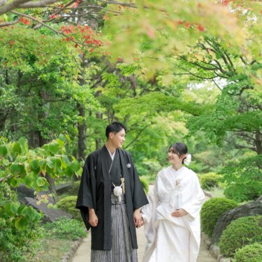 Bride in shiromuku and groom in montsuki and hakama walking hand in hand along a garden path framed by green and red maple leaves, a natural kimono wedding photoshoot moment