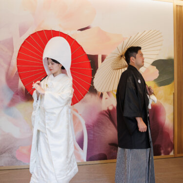 Bride in wataboshi with a red wagasa umbrella and groom with a white one stand apart before a pastel floral mural, a Japanese studio wedding photoshoot