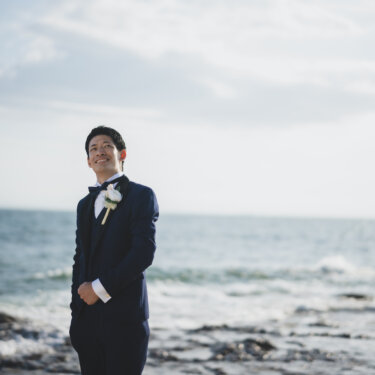 Groom in a navy tuxedo with a white boutonniere laughing by the waves, a solo portrait from a beach pre-wedding photoshoot on the Chita Peninsula