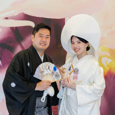 Bride in shiromuku and wataboshi and groom in montsuki hold folding fans reading 'husband' and 'wife' in front of a floral mural, kimono wedding photos
