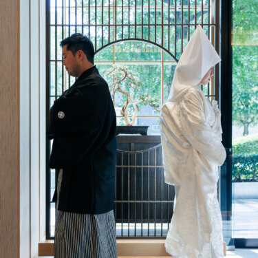 Groom in montsuki hakama and bride in a pointed wataboshi hood stand back to back by a lattice screen, a stylish Japanese wedding photoshoot pose