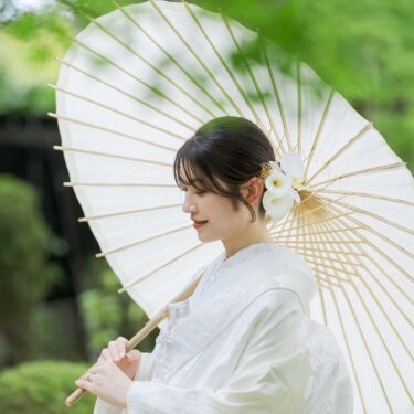 Bride in a white shiromuku with an orchid hair ornament holding a white wagasa umbrella under fresh green maples at Daisen Park, a graceful pre-wedding portrait in Osaka
