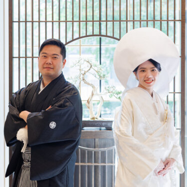 Groom with arms crossed in montsuki and bride in shiromuku and wataboshi stand before a lattice partition with an arched window, kimono wedding photos