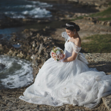 Bride sitting on the rocky shore in a voluminous ball gown wedding dress, gazing at her bouquet beside the crashing waves in her wedding photos