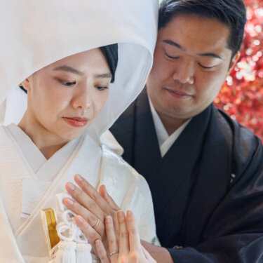 Close-up of the bride in wataboshi showing her ring as the groom in montsuki leans in from behind, red maple leaves behind them in a kimono wedding photo