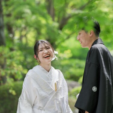 Bride in shiromuku laughing with the groom in black montsuki among lush garden greenery, a natural candid moment from their kimono wedding photos