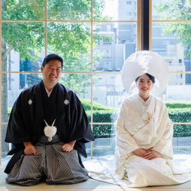 Groom in montsuki hakama and bride in shiromuku and wataboshi sit formally on tatami in front of a window with city greenery, Japan wedding photography