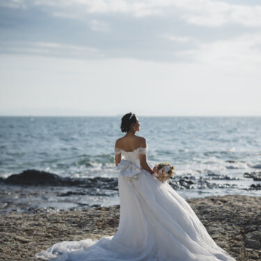 Back view of the bride standing on the rocks with her long wedding dress train spread out toward the open sea, a classic beach wedding photography shot