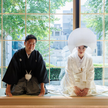 Groom in montsuki hakama and bride in shiromuku with a wataboshi bow politely side by side on tatami by a large window, a Japanese wedding photoshoot