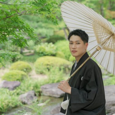 Groom in black montsuki holding a white paper umbrella over his shoulder beside the pond and rocks of Daisen Park Japanese Garden, a classic Japan wedding photography portrait