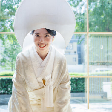 Bride wearing a round white wataboshi hood and shiromuku kneels on tatami and smiles at the camera, a traditional Japanese wedding photo