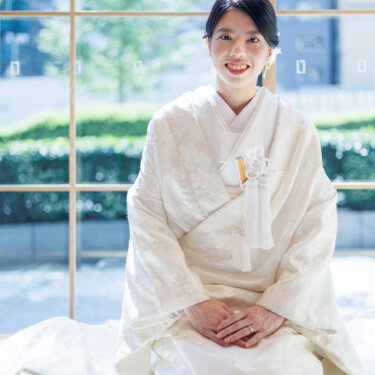 Smiling bride in shiromuku kneels on tatami with her hands folded, green trees visible through the window behind her, kimono wedding photography