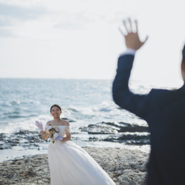 Bride waving with a bouquet toward the groom on the rocky seaside, a candid moment from a pre-wedding photoshoot in Aichi