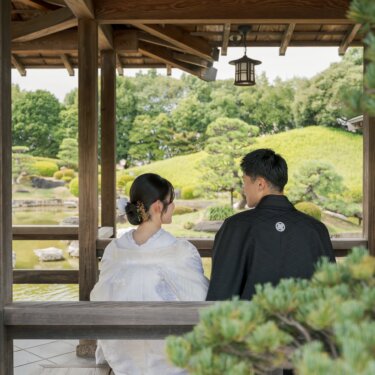 Couple in kimono seen from behind, sitting under the eaves of a garden pavilion and gazing over the pond and pine trees, a calm back-view pose for wedding photos in Japan
