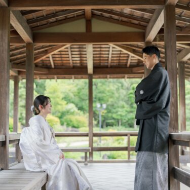 Bride in shiromuku seated on a pavilion bench while the groom in montsuki leans on the railing and smiles at her, a relaxed kimono wedding photography scene at Daisen Park