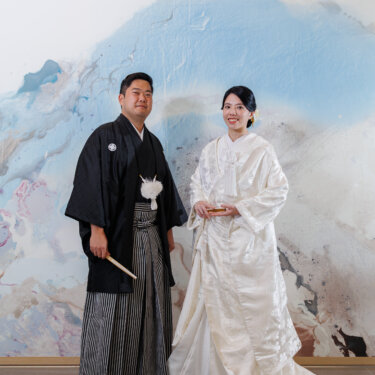 Groom in montsuki hakama and bride in shiromuku stand side by side before a pastel abstract mural, a full-length kimono wedding photo in Japan
