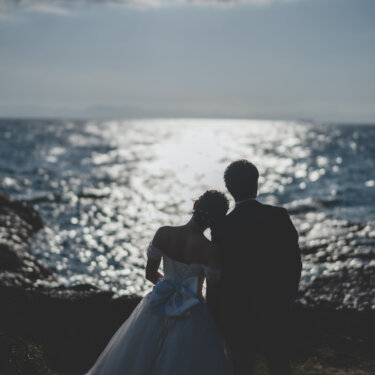 Bride and groom standing close on the dark rocks watching the sun glitter on the sea, a quiet back view from their wedding photoshoot on the Chita Peninsula