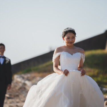 Bride lifting the skirt of her off-shoulder wedding dress as the groom watches from behind, a playful first look style pose for beach wedding photos