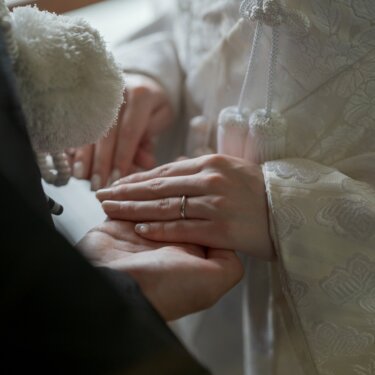 Close-up of the bride's hand with her wedding ring resting on the groom's palm, framed by white tassels and the silk of her shiromuku, a detail shot from a kimono wedding photoshoot