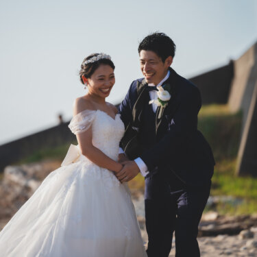 Laughing bride and groom holding hands on the shore by a concrete seawall, natural smiles captured in pre-wedding photography in Japan