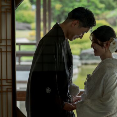 Bride and groom in shiromuku and montsuki touching foreheads by a wooden lattice in a pavilion at Daisen Park, softly backlit against the pond in this pre-wedding photo