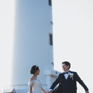 Groom leading the bride by the hand in front of the white tower of Noma Lighthouse, a joyful wedding photoshoot moment in Aichi