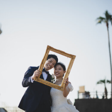 Couple peeking through an antique gold frame beside Noma Lighthouse and palm trees, a creative prop idea for wedding photography
