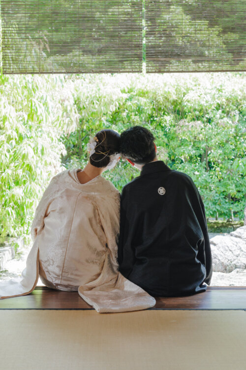 Back view of a bride in shiromuku and a groom in montsuki sitting side by side on a veranda, looking out at a green garden through bamboo blinds in a kimono wedding photoshoot in Japan