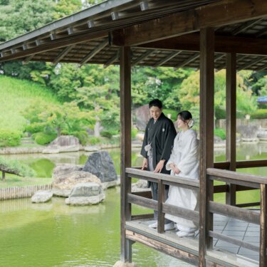 Couple in kimono leaning on the railing of a waterside pavilion above the pond at Daisen Park Japanese Garden, with a red-pillared pavilion across the water, a scenic wedding photography shot in Osaka