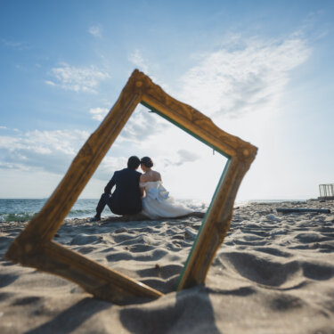 Gold picture frame standing in the sand framing the couple sitting by the sea with the lighthouse behind, a creative beach wedding photo composition