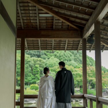 Back view of the bride in shiromuku and the groom in montsuki and hakama standing at the end of a covered garden corridor, looking out over green hills for their pre-wedding photos