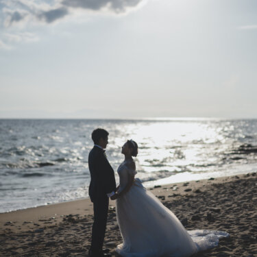 Bride and groom holding hands face to face on the sandy beach as the sea sparkles in the afternoon light, a romantic Japan wedding photography scene