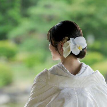 Back view of the bride's elegant low updo adorned with a white orchid and gold kanzashi, the shiromuku collar framed by the soft green garden, a bridal hairstyle for kimono wedding photos