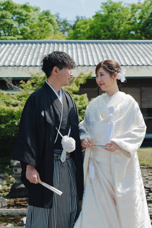 Groom in montsuki hakama holding a fan and bride in shiromuku gazing at each other before a tiled-roof building in a Japanese garden, kimono wedding photos in Japan