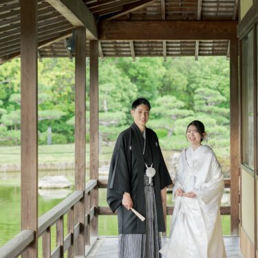 Couple in shiromuku and striped hakama standing together in the long wooden corridor of Daisen Park Japanese Garden, the groom holding a folding fan, for their kimono wedding photos