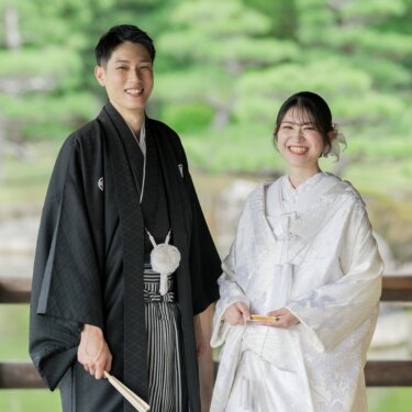 Groom in montsuki holding a folding fan and bride in shiromuku laughing together in front of a pine-lined pond, a joyful full-length kimono wedding photography portrait