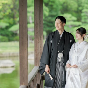 Groom and bride in montsuki and shiromuku smiling side by side between the wooden pillars of a pavilion at Daisen Park, a natural pose for pre-wedding photos in Osaka
