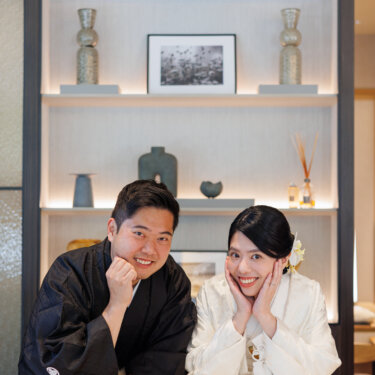 Bride in shiromuku and groom in montsuki lean on a counter with their chins in their hands and big smiles, a playful kimono wedding photos pose