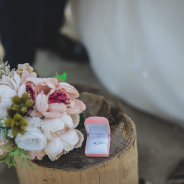 Bridal bouquet of pink peonies and a pink ring box placed on a driftwood stump in the sand, wedding photo props from a beach photoshoot