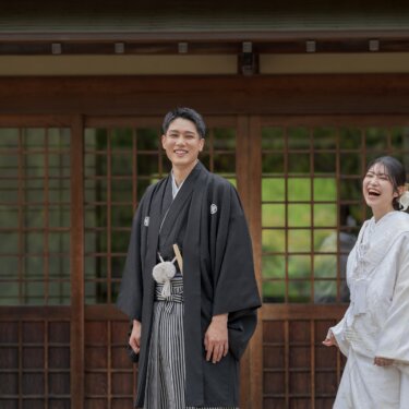 Groom in montsuki and bride in shiromuku laughing in front of the latticed wooden doors of a traditional Japanese house, a cheerful moment from their wedding photoshoot